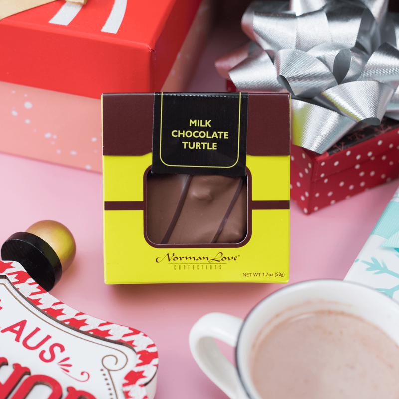 A Milk Chocolate Turtle sitting upright in a green box on a pink background. Gift boxes and a mug of hot chocolate bring forth the spirit of the season. 
